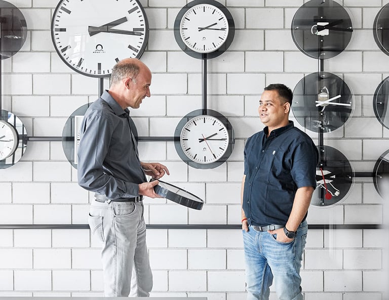 Two people from MOBATIME discuss the topic of GNSS Two people are standing in front of a wall with various clocks and discussing about GNSS
